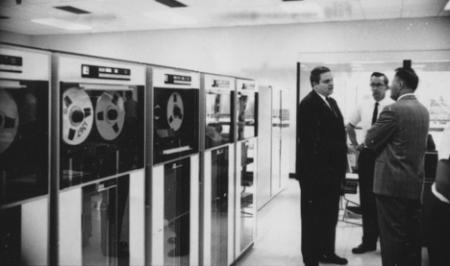Three men conversing in front of an old data-processing room that uses tape.